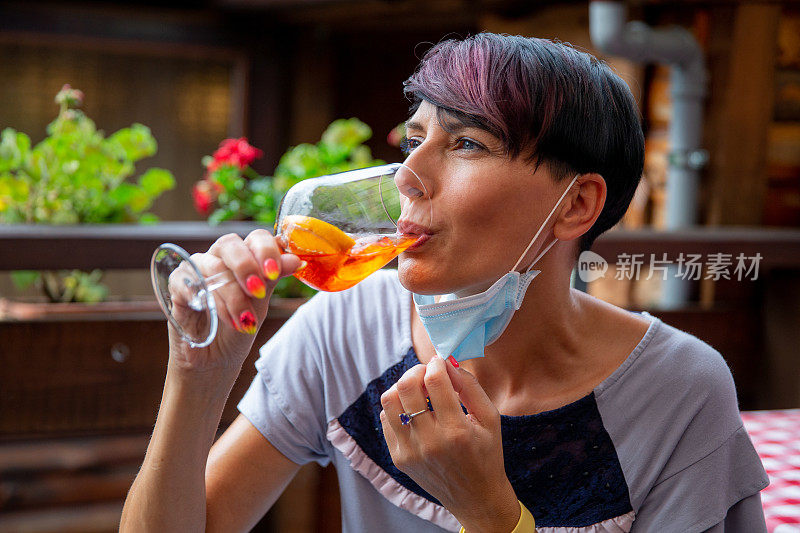 Mid Adult Woman Holding Her Protective Face Mask Down While Drinking in Bar .中年妇女在酒吧里喝酒时戴着保护面罩